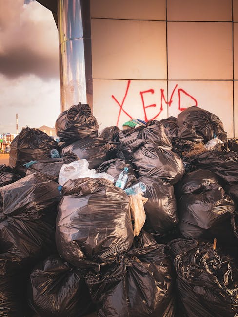 A large pile of black plastic rubbish bags, predominantly tied at the tops, is stacked against a reflective metal wall on a paved or concrete surface, likely an urban outdoor location. The bags contai