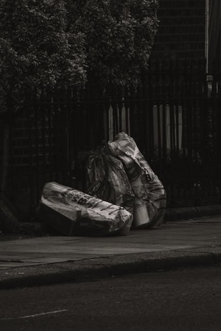 A collection of overflowing rubbish bins and scattered waste on a paved urban street corner in front of a commercial building with storefronts. The waste includes paper, cardboard boxes, plastic bags,