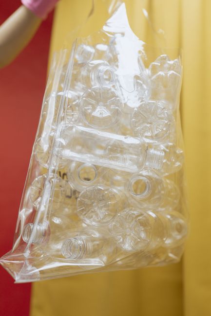 A clear plastic bag filled with transparent empty plastic bottles, flutes, and caps, held by a person wearing a pink sleeve against a background of red and yellow fabric or wall. The bottles vary in s