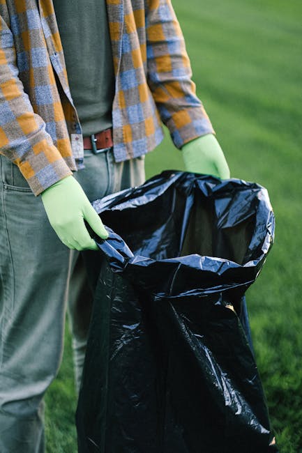 A pile of mixed waste materials is situated against a brick and concrete wall outdoors. The waste includes flattened cardboard boxes, some with visible printed barcode labels, partially crushed and fo